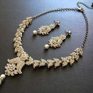 Op de afbeelding: Een zilverkleurige ketting- en oorbellenset met heldere kristallen strass-steentjes in bladvormige patronen. De ketting heeft een centrale hanger met een bungelende witte kunstparel, en de bijpassende oorbellen bevatten eveneens witte kunstparels.