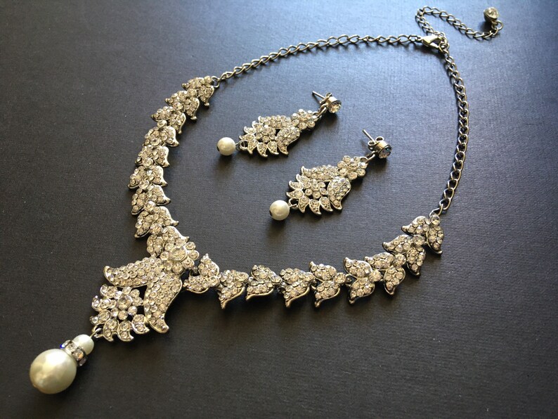 Op de afbeelding: Een zilverkleurige ketting- en oorbellenset met heldere kristallen strass-steentjes in bladvormige patronen. De ketting heeft een centrale hanger met een bungelende witte kunstparel, en de bijpassende oorbellen bevatten eveneens witte kunstparels.