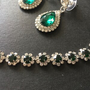 May include: A jewellery set featuring a necklace and drop earrings. The necklace consists of circular links with dark green round stones surrounded by clear crystals. The earrings feature a large pear-shaped dark green stone with a double halo of clear crystals.