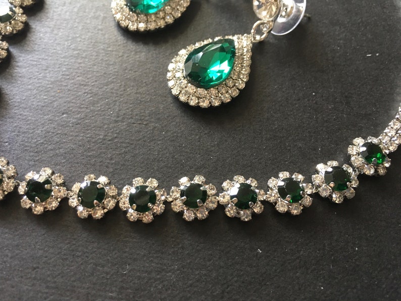 May include: A jewellery set featuring a necklace and drop earrings. The necklace consists of circular links with dark green round stones surrounded by clear crystals. The earrings feature a large pear-shaped dark green stone with a double halo of clear crystals.