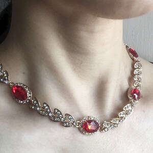 May include: A gold necklace with a delicate leaf design and red gemstone accents. The necklace features a larger oval-shaped red gemstone in the center.