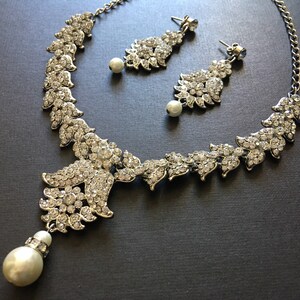 Op de afbeelding: Een zilverkleurige set van een ketting en oorbellen met doorzichtige kristallen strass-steentjes in bladvormige patronen. De ketting heeft een centraal bloemmotief met een bungelende witte kunstparel, en elke oorbel is voorzien van een parel.