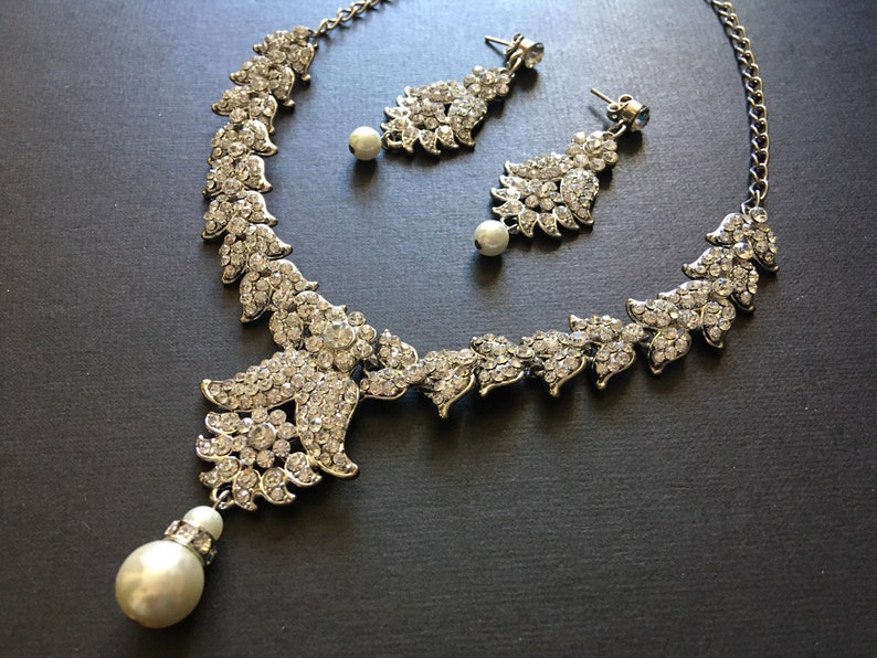 Op de afbeelding: Een zilverkleurige set van een ketting en oorbellen met doorzichtige kristallen strass-steentjes in bladvormige patronen. De ketting heeft een centraal bloemmotief met een bungelende witte kunstparel, en elke oorbel is voorzien van een parel.