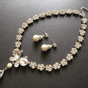 Puede incluir: Un conjunto de collar y pendientes plateados. El collar presenta un dise&ntilde;o floral con cristales transparentes y un gran colgante de cristal en forma de l&aacute;grima. Los pendientes tienen perlas. Las joyas se exhiben sobre una superficie oscura.