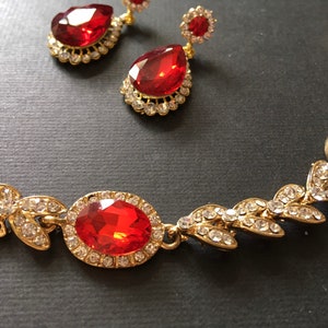 May include: A gold necklace with a red gemstone pendant and leaf-shaped accents. The necklace is adorned with clear rhinestones.