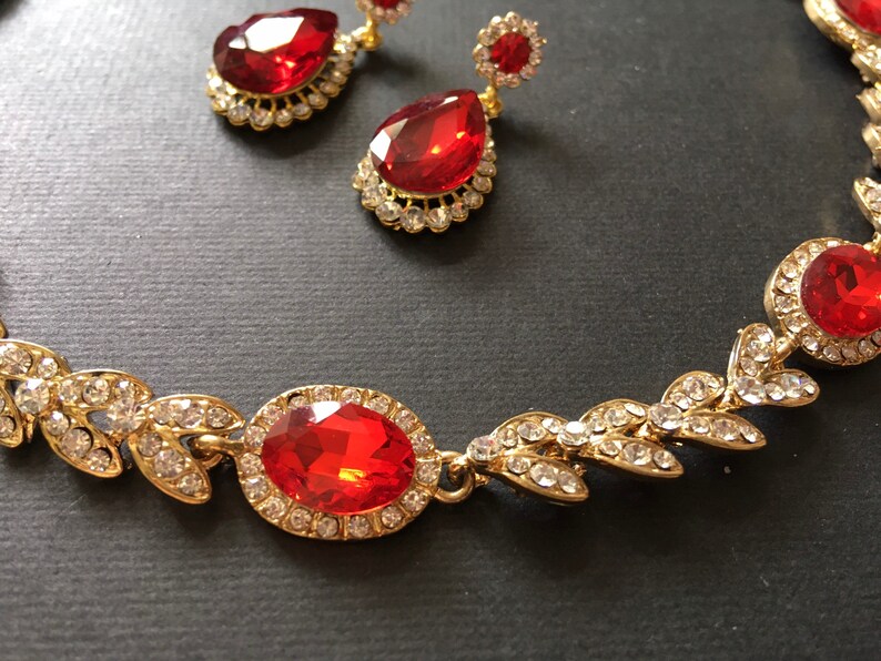 May include: A gold necklace with a red gemstone pendant and leaf-shaped accents. The necklace is adorned with clear rhinestones.