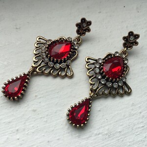 Könnte beinhalten: Ein Paar verzierter Ohrhänger im Vintage-Stil. Jeder Ohrring hat einen großen, tropfenförmigen roten Edelstein, der in einer filigranen Fassung mit kleineren, klaren Steinen und einem floralen Akzent gefasst ist. Das Metall hat eine bronzefarbene Oberfläche.