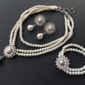 Può includere: Un set di gioielli composto da una collana di perle a tre fili, orecchini abbinati e un braccialetto a due fili. Ogni pezzo presenta un motivo floreale argentato con una perla centrale e cristalli trasparenti. Il set è su una superficie grigio scuro.