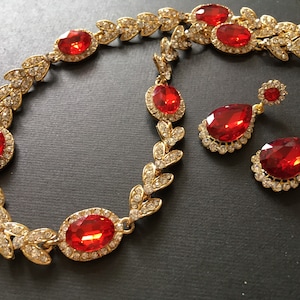 May include: A gold necklace and earrings set with red and clear rhinestones. The necklace features a leaf design and the earrings are teardrop shaped.