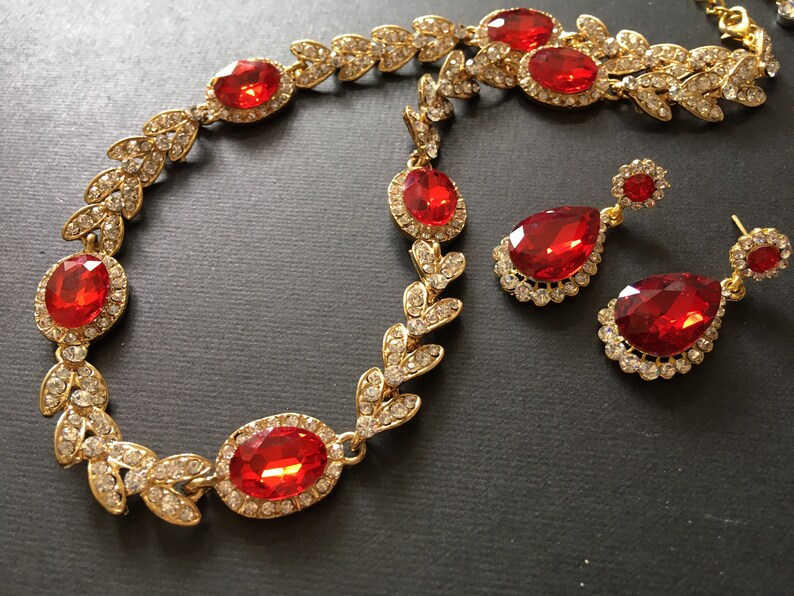 May include: A gold necklace and earrings set with red and clear rhinestones. The necklace features a leaf design and the earrings are teardrop shaped.