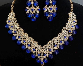 May include: A gold-tone necklace and matching drop earring set featuring intricate filigree metalwork, clear white rhinestones, and round, faceted royal blue crystals. The necklace has a V-shaped design with a central dangling blue crystal.