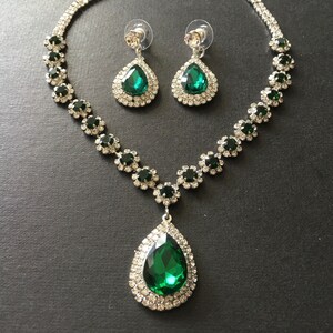 May include: A jewellery set consisting of a necklace and matching drop earrings. The necklace features a large, pear-shaped emerald green stone pendant surrounded by clear crystals, with a chain of alternating green and clear crystal floral links.