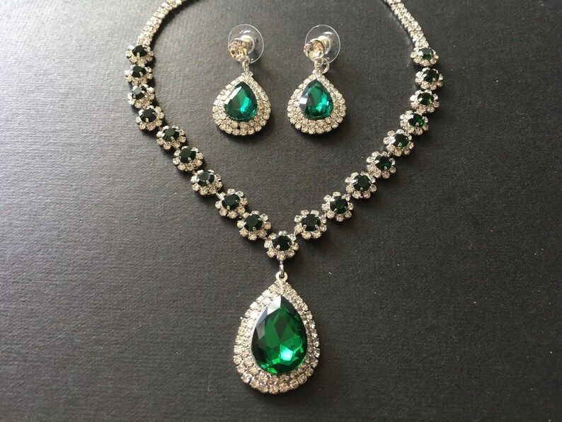 May include: A jewellery set consisting of a necklace and matching drop earrings. The necklace features a large, pear-shaped emerald green stone pendant surrounded by clear crystals, with a chain of alternating green and clear crystal floral links.