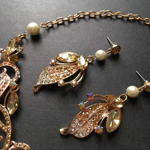 May include: Gold-toned jewelry set featuring a necklace and earrings. The necklace and earrings have a leaf design adorned with clear and amber-colored crystals. The earrings include a pearl drop. The jewelry is set against a dark background.