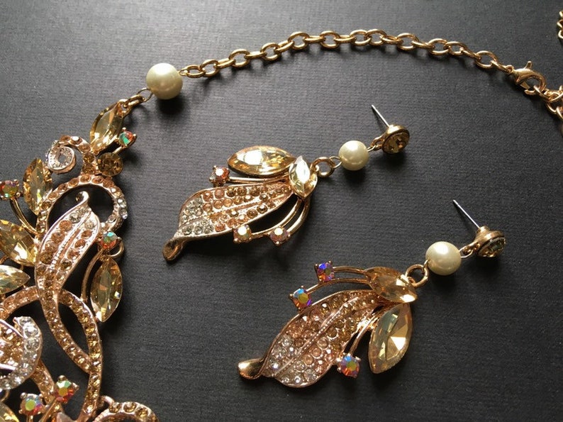 May include: Gold-toned jewelry set featuring a necklace and earrings. The necklace and earrings have a leaf design adorned with clear and amber-colored crystals. The earrings include a pearl drop. The jewelry is set against a dark background.