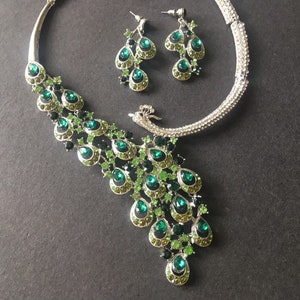 May include: A silver peacock-shaped necklace and earrings set with green and black rhinestones. The necklace has a choker style with a peacock tail design.