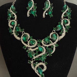 May include: A jewelry set consisting of a necklace and matching earrings featuring large emerald green faceted crystals set in silver-toned metal with smaller clear rhinestones. The necklace has a swirling, asymmetrical design with the text Weddingvalle.