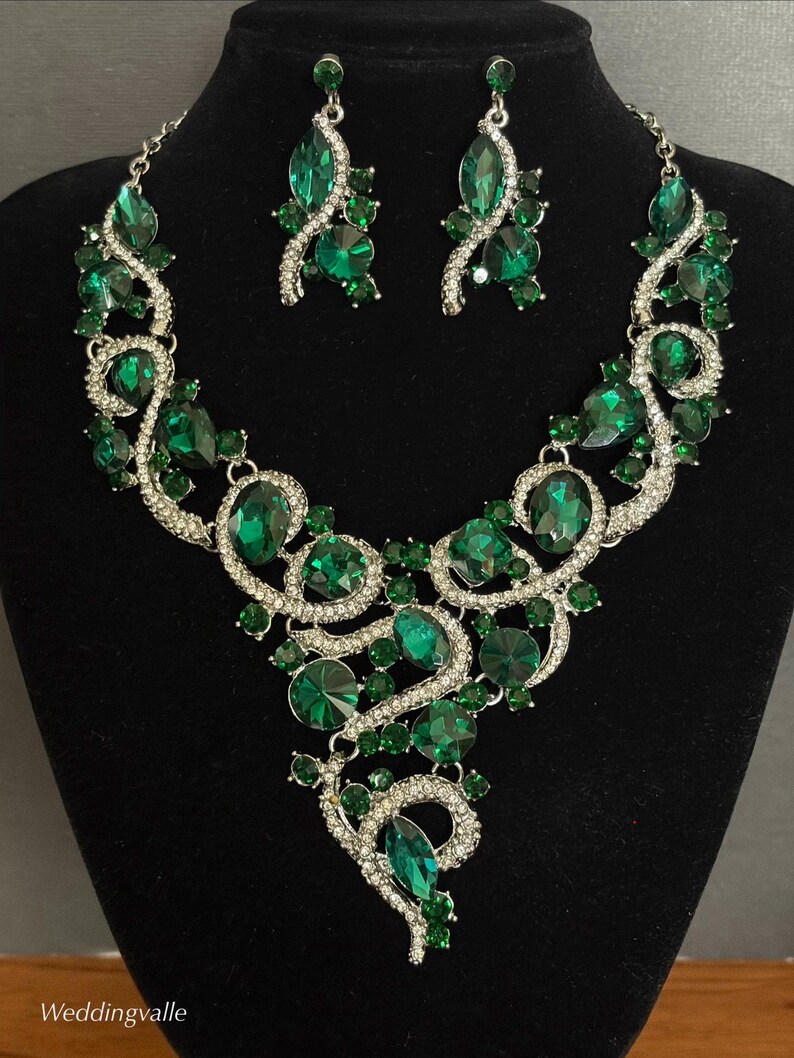May include: A jewelry set consisting of a necklace and matching earrings featuring large emerald green faceted crystals set in silver-toned metal with smaller clear rhinestones. The necklace has a swirling, asymmetrical design with the text Weddingvalle.