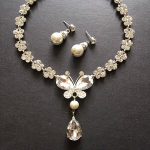 Puede incluir: Un conjunto de collar y pendientes plateados. El collar presenta un colgante de mariposa con cristales transparentes y un cristal en forma de l&aacute;grima. Los pendientes tienen perlas colgantes. El collar tiene un dise&ntilde;o floral.