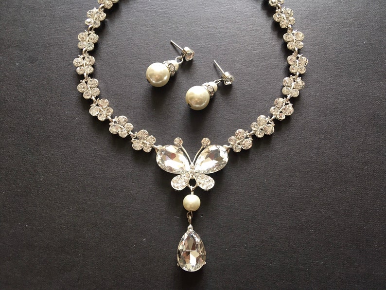 Puede incluir: Un conjunto de collar y pendientes plateados. El collar presenta un colgante de mariposa con cristales transparentes y un cristal en forma de l&aacute;grima. Los pendientes tienen perlas colgantes. El collar tiene un dise&ntilde;o floral.