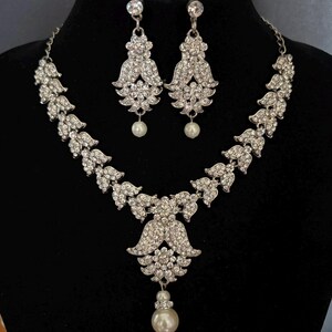 Op de afbeelding: Een zilverkleurige sieradenset bestaande uit een ketting en bijpassende oorbellen. De ketting heeft een bladpatroon versierd met heldere kristallen en een centrale hanger met een witte parel. Elke oorbel heeft een met kristallen ingelegd bloemmotief met een bungelende witte parel.