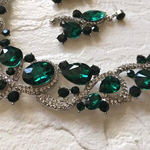May include: A silver-toned necklace and earring set featuring large emerald green faceted crystals, smaller round black stones, and clear pav&eacute; rhinestones set in a swirling metal frame. The jewelry pieces display a mix of oval, pear, and round shapes.