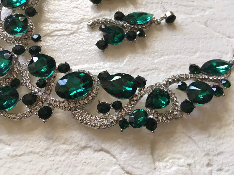 May include: A silver-toned necklace and earring set featuring large emerald green faceted crystals, smaller round black stones, and clear pav&eacute; rhinestones set in a swirling metal frame. The jewelry pieces display a mix of oval, pear, and round shapes.