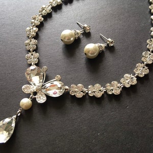 Puede incluir: Un conjunto de joyas plateadas con collar y pendientes a juego. El collar presenta un dise&ntilde;o floral con cristales transparentes y un gran colgante en forma de l&aacute;grima. Los pendientes son pendientes de perlas con detalles de cristal. El conjunto se muestra sobre una superficie oscura.
