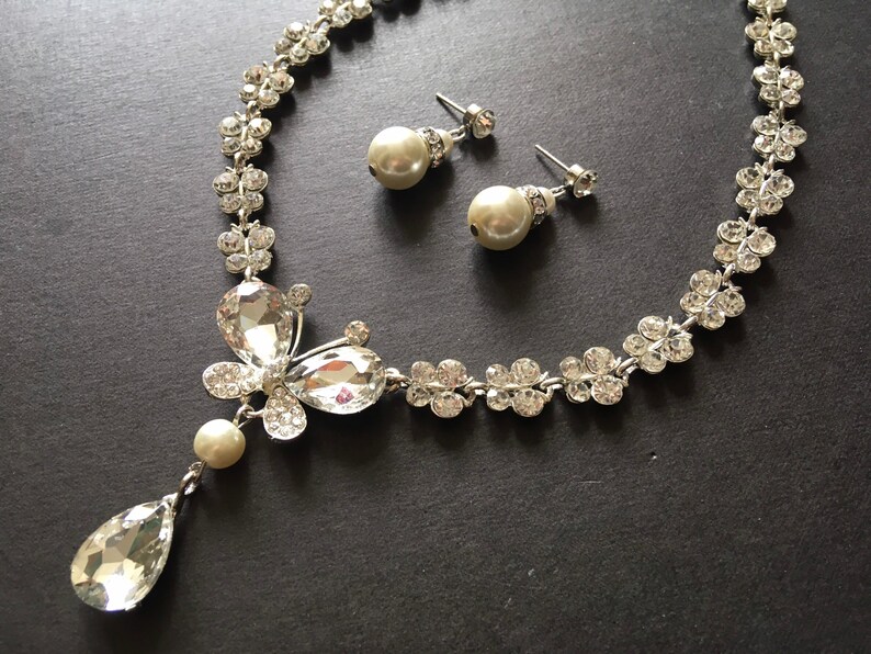 Puede incluir: Un conjunto de joyas plateadas con collar y pendientes a juego. El collar presenta un dise&ntilde;o floral con cristales transparentes y un gran colgante en forma de l&aacute;grima. Los pendientes son pendientes de perlas con detalles de cristal. El conjunto se muestra sobre una superficie oscura.