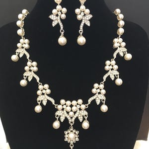 May include: A silver-toned necklace and earring set with faux pearls and clear rhinestones. The necklace features a floral design with dangling pearls, and the earrings have a similar style. The jewelry is displayed on a black mannequin.
