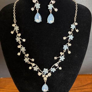Può includere: Un set di gioielli color argento composto da una collana e orecchini pendenti abbinati. La collana presenta un design a tralcio con accenti floreali azzurri e cristalli trasparenti, terminando con un ciondolo azzurro a forma di goccia.