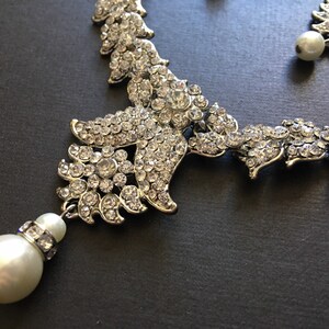 Op de afbeelding: Een zilverkleurige ketting met ingewikkelde bloemvormige metalen schakels ingelegd met heldere, sprankelende strass-steentjes. Het ontwerp bevat een bungelende witte kunstparel in het midden, geaccentueerd door een klein bandje strass-steentjes.