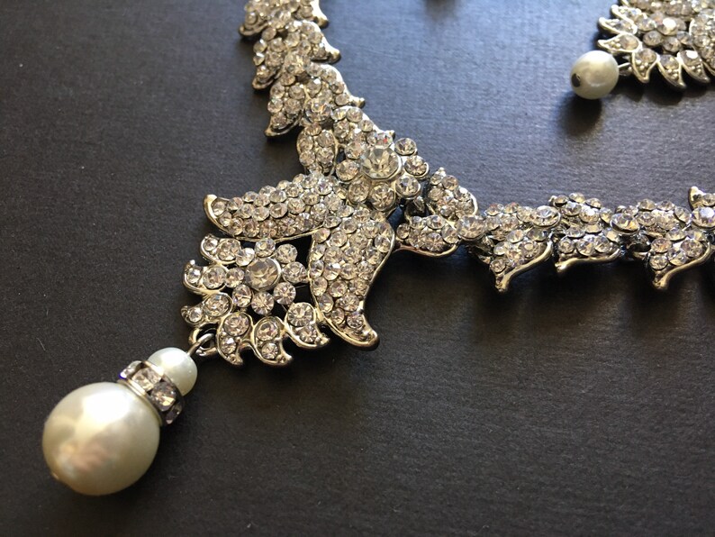 Op de afbeelding: Een zilverkleurige ketting met ingewikkelde bloemvormige metalen schakels ingelegd met heldere, sprankelende strass-steentjes. Het ontwerp bevat een bungelende witte kunstparel in het midden, geaccentueerd door een klein bandje strass-steentjes.