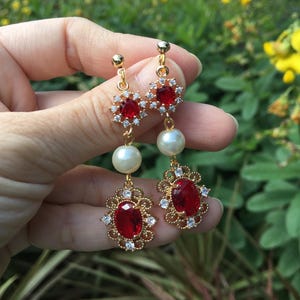 Może przedstawiać: Para złotych kolczyków z czerwonymi i białymi kamieniami. Kolczyki mają duży czerwony kamień otoczony mniejszymi białymi kamieniami i mniejszy czerwony kamień otoczony mniejszymi białymi kamieniami. Kolczyki wiszą na złotym łańcuszku z perłą.