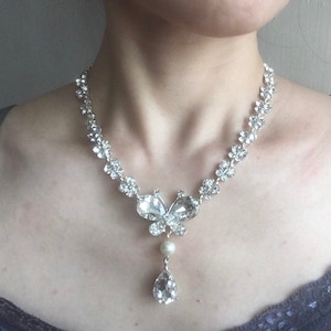 Puede incluir: Collar plateado con un colgante de mariposa con cristales transparentes y una gota de cristal en forma de l&aacute;grima. La cadena presenta un dise&ntilde;o floral. El collar incluye una perla. Una joya delicada.