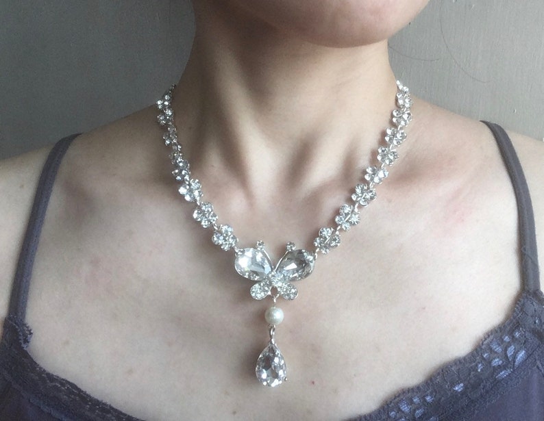 Puede incluir: Collar plateado con un colgante de mariposa con cristales transparentes y una gota de cristal en forma de l&aacute;grima. La cadena presenta un dise&ntilde;o floral. El collar incluye una perla. Una joya delicada.