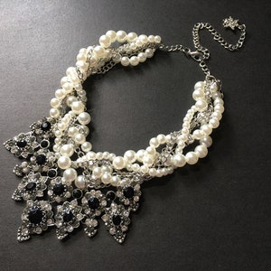Puede incluir: Un collar de perlas de múltiples hilos con un gran colgante adornado con pedrería negra y plateada. El colgante tiene forma de lágrima y termina en punta.