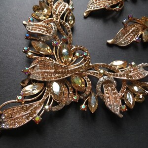 May include: A gold-toned jewelry set featuring a necklace and earrings. The necklace and earrings have a floral design with clear and amber-colored crystals. The set is on a dark background.