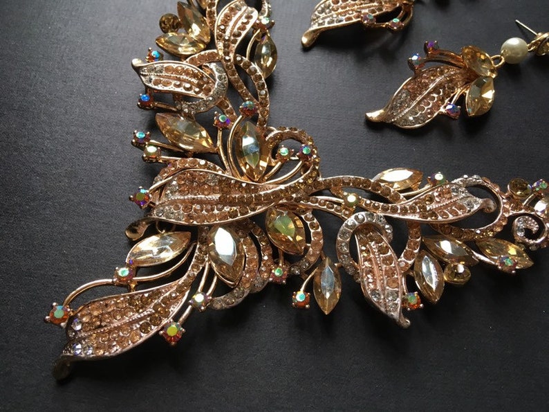 May include: A gold-toned jewelry set featuring a necklace and earrings. The necklace and earrings have a floral design with clear and amber-colored crystals. The set is on a dark background.