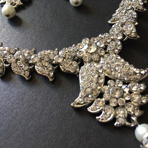 Op de afbeelding: Een zilverkleurige ketting- en oorbellenset met heldere, gefacetteerde strass-steentjes in een bladvormig patroon. De sieraden zijn versierd met ronde, witte kunstparels, wat zorgt voor een bijpassende set met een metallic afwerking en fonkelende stenen.
