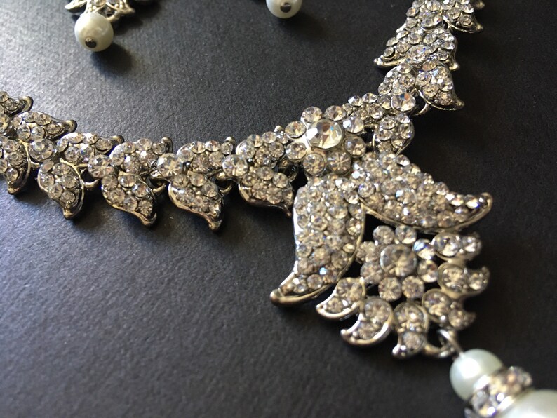 Op de afbeelding: Een zilverkleurige ketting- en oorbellenset met heldere, gefacetteerde strass-steentjes in een bladvormig patroon. De sieraden zijn versierd met ronde, witte kunstparels, wat zorgt voor een bijpassende set met een metallic afwerking en fonkelende stenen.
