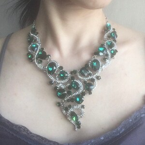 May include: A statement necklace featuring a silver-toned metal base adorned with numerous dark emerald green faceted crystals and small clear rhinestones. The design consists of swirling, interconnected metal links that form a V-shaped pattern.