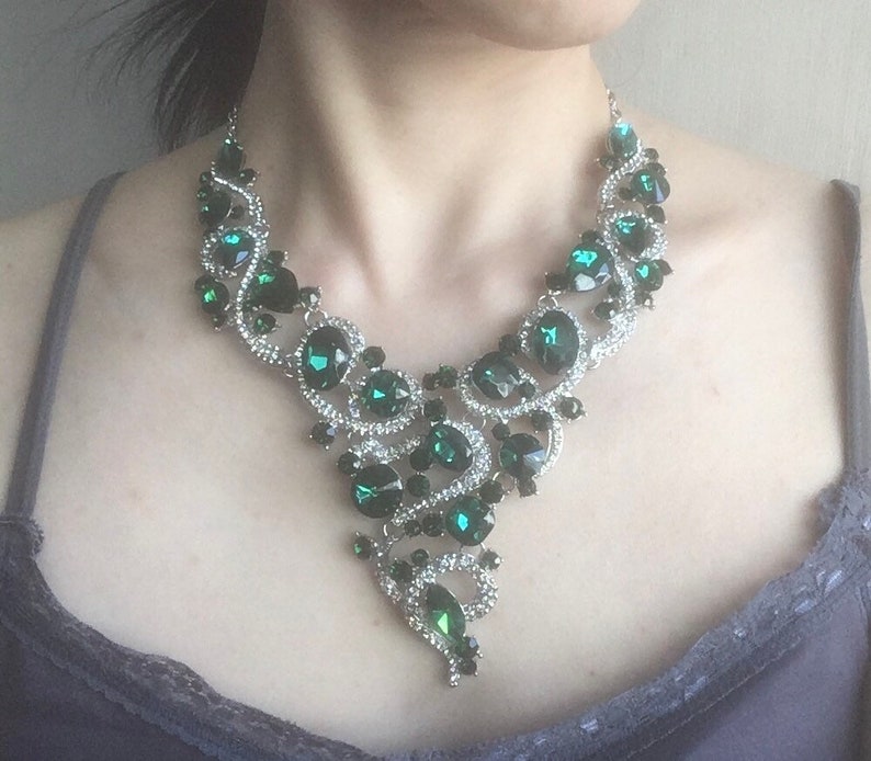 May include: A statement necklace featuring a silver-toned metal base adorned with numerous dark emerald green faceted crystals and small clear rhinestones. The design consists of swirling, interconnected metal links that form a V-shaped pattern.