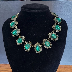May include: A statement necklace featuring a series of oval-shaped emerald green faceted stones set in antique gold-toned metal frames. Each large central stone is surrounded by smaller green crystals, connected by metal links. Text reads Weddingvalle.