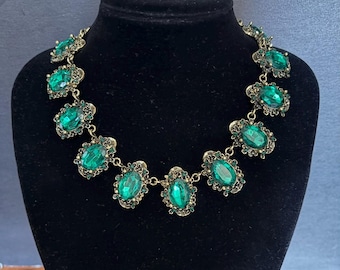 Op de afbeelding: Een statement ketting met een reeks ovaalvormige smaragdgroene gefacetteerde stenen in antiek goudkleurige metalen monturen. Elke grote centrale steen is omringd door kleinere groene kristallen, verbonden door metalen schakels. Tekst leest Weddingvalle.