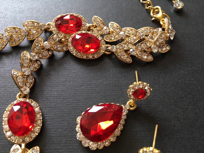 May include: A gold-tone necklace with red and clear crystal stones. The necklace features a leaf design and is paired with matching earrings.