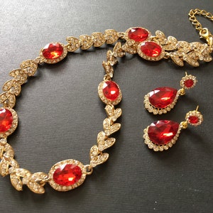 May include: A gold necklace with red gemstone accents and matching earrings. The necklace features a leaf design with a large red gemstone in the center. The earrings are teardrop shaped with a red gemstone in the center.