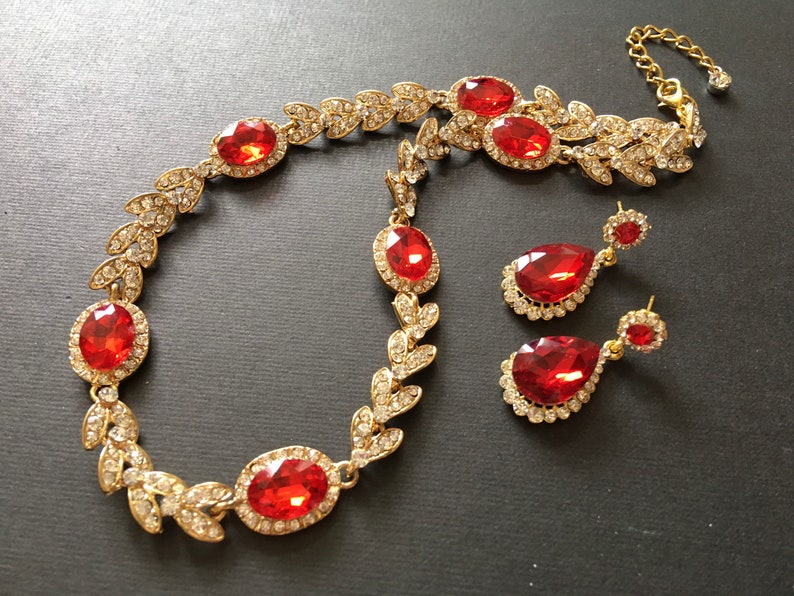 May include: A gold necklace with red gemstone accents and matching earrings. The necklace features a leaf design with a large red gemstone in the center. The earrings are teardrop shaped with a red gemstone in the center.