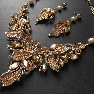 May include: A gold-toned jewelry set featuring a necklace and matching earrings. The necklace has a floral design with clear and amber-colored crystals. The earrings have a leaf design with pearl accents. The set is on a black background.