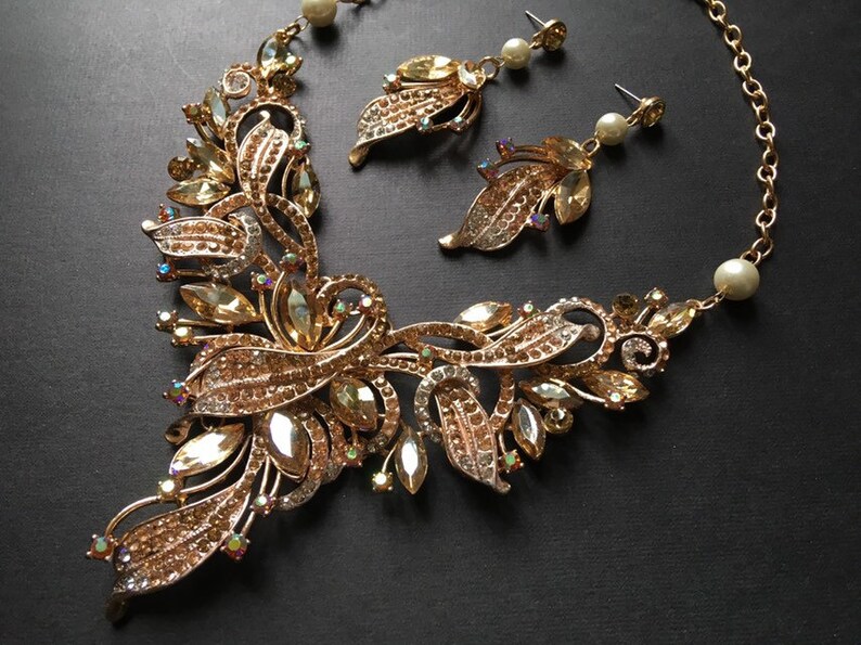 May include: A gold-toned jewelry set featuring a necklace and matching earrings. The necklace has a floral design with clear and amber-colored crystals. The earrings have a leaf design with pearl accents. The set is on a black background.